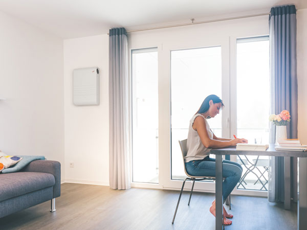 Innenraum mit großer Fensterfront, Tisch und Lüftungsgerät an der Wand.