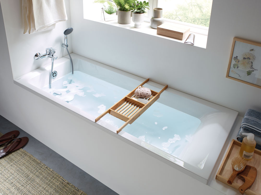Helle Badewanne aus der Geberit Renova Plan Serie mit Wandarmatur und Holzablage.